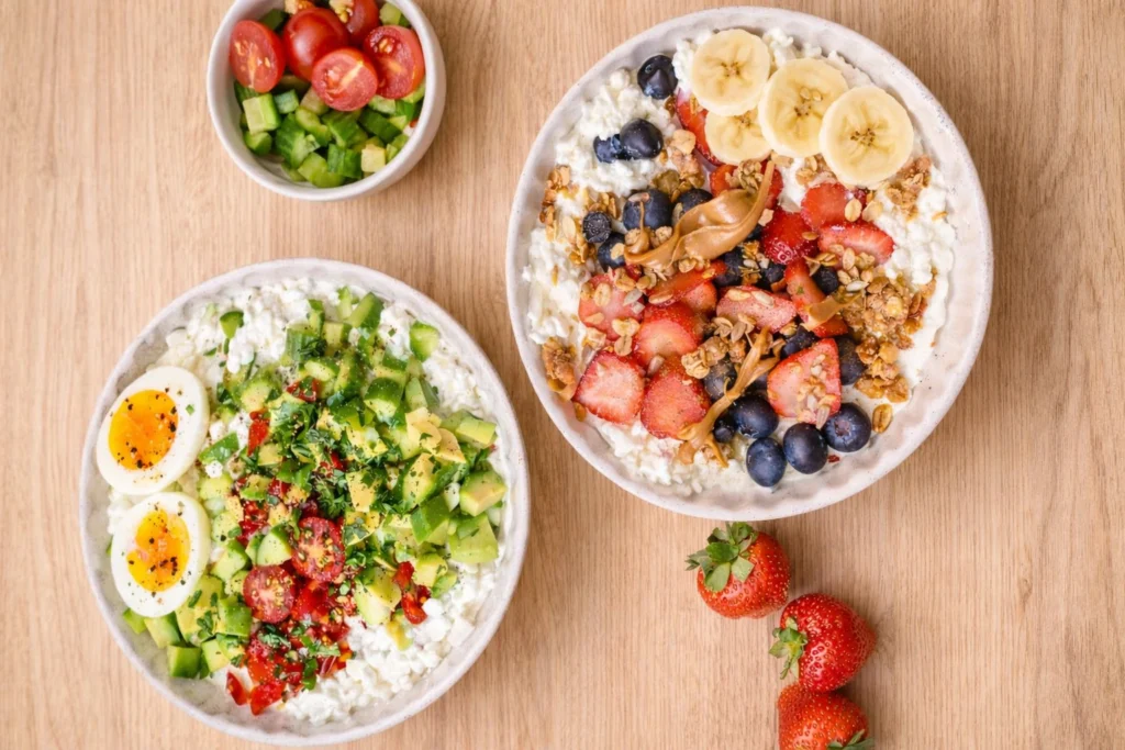 Cottage Cheese Breakfast Bowl sweet and savory with fruits, granola, banana, avocado, cucumber and eggs on a beige wooden surface.