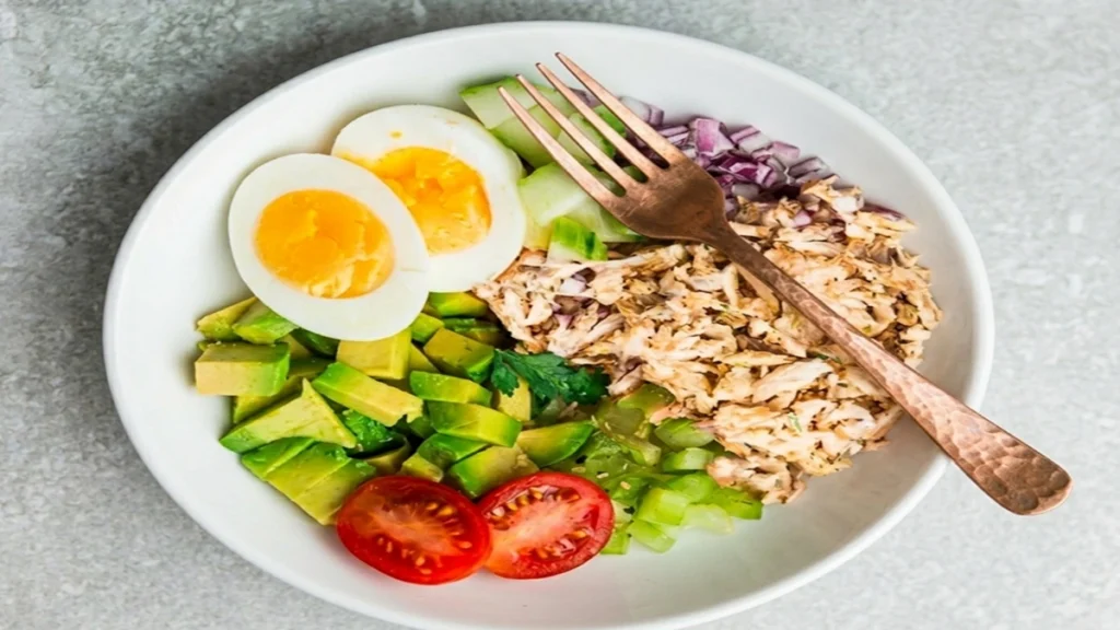 Healthy Keto Avocado Chicken Salad with boiled eggs, fresh avocado, shredded chicken, cucumber, and tomatoes in a white bowl.