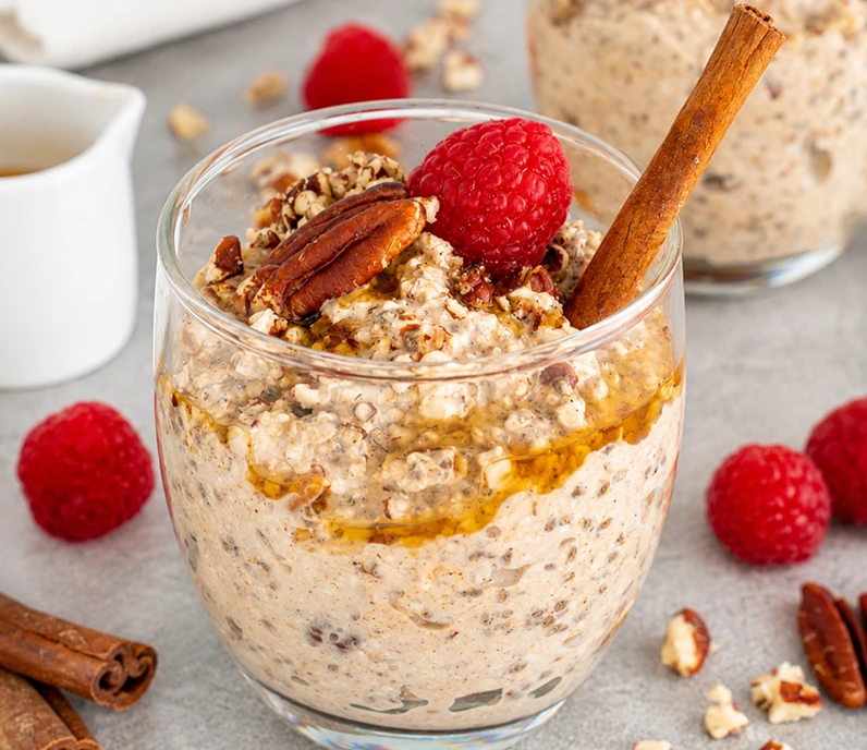 Creamy Maple Pecan Overnight Oats topped with pecans, raspberries, cinnamon sticks, and maple drizzle in a glass jar.
