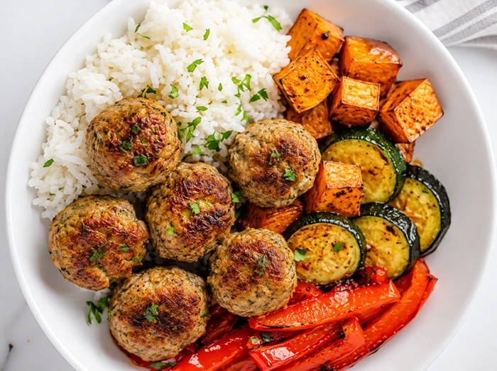 Meatballs with Roasted Veggies & Rice served in a white bowl with roasted zucchini, sweet potatoes, red peppers, and fluffy white rice.