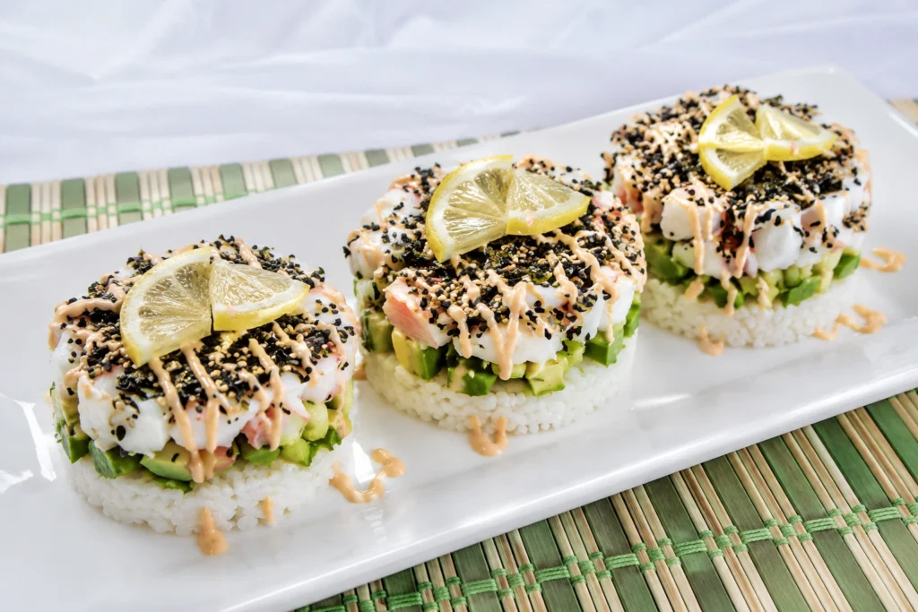 Three California Sushi Stack portions served on a white plate with avocado, crab, sesame seeds, spicy mayo drizzle, and lemon slices.