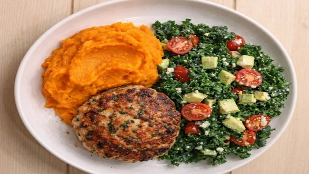 A white bowl featuring a Sweet Potato and Turkey Plate with a seared turkey patty, a scoop of mashed sweet potato, and a chopped kale salad topped with avocado, cherry tomatoes, and feta cheese, served on a light wood surface.