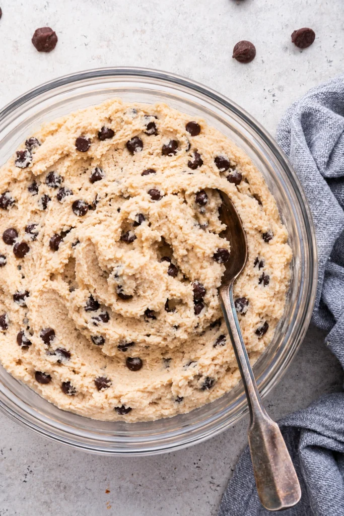 Creamy Cottage Cheese Cookie Dough in a clear glass bowl topped with mini chocolate chips, photographed from above with a spoon on a light background.
