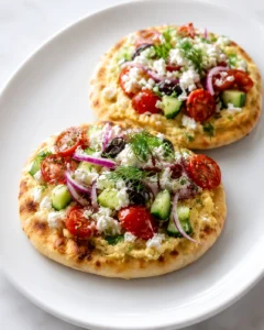 Mediterranean Mini Lahmacun Bites with Yogurt Drizzle topped with cherry tomatoes, cucumber, red onion, feta cheese, olives, and fresh herbs served on a white plate.