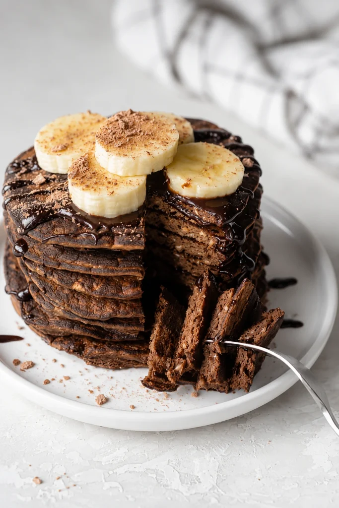 Double Chocolate Protein Waffles stacked high on a white round plate, topped with banana slices and rich chocolate drizzle, creating a high-protein chocolate breakfast.