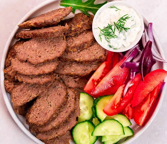 Easy Homemade Gyro Meat recipe served in a bowl with sliced gyro meat, fresh tomatoes, cucumbers, red onions, and creamy tzatziki sauce.