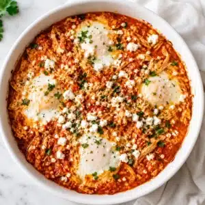 Mediterranean Egg Bake served in a white round baking dish with baked eggs, feta cheese, shredded chicken, tomato sauce, and fresh parsley for a high-protein breakfast.