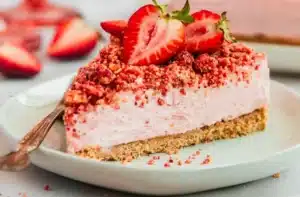 Strawberry Crunch Cheesecake Fluff slice with a buttery graham cracker crust, creamy strawberry cheesecake filling, crunchy strawberry topping, and fresh strawberries served on a white round plate.