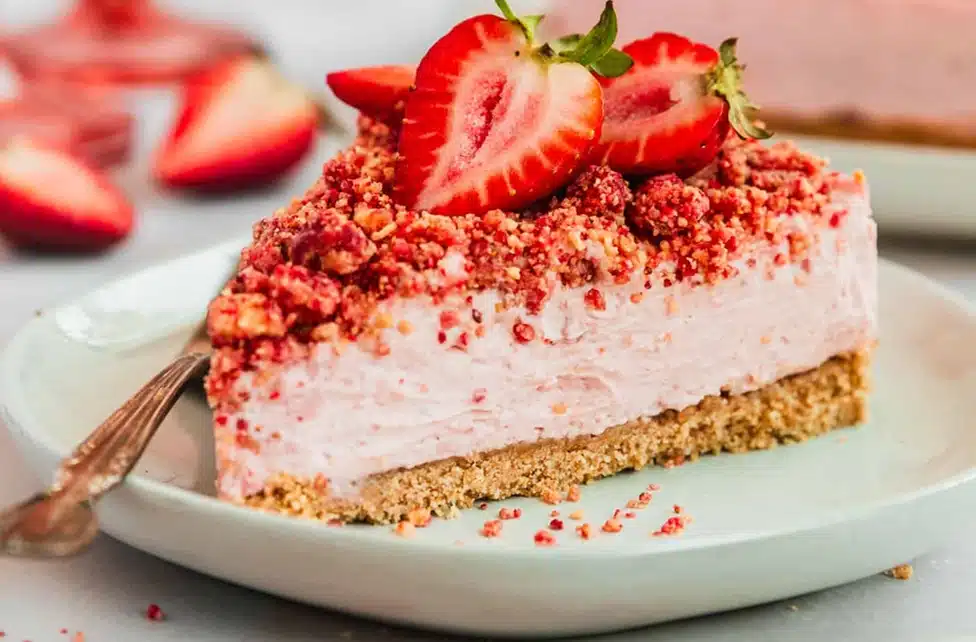 Strawberry Crunch Cheesecake Fluff slice with a buttery graham cracker crust, creamy strawberry cheesecake filling, crunchy strawberry topping, and fresh strawberries served on a white round plate.