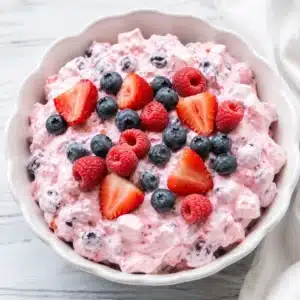 Triple Berry Fluff Dessert served in a white scalloped bowl, topped with fresh strawberries, blueberries, and raspberries for a creamy no-bake summer dessert.